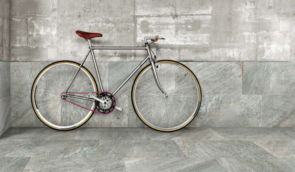 La collezione Discovery Colonial, nel formato 30x60, caratterizza questo spazio moderno con piastrelle in gres porcellanato effetto pietra dalle eleganti sfumature grigio-verdi. Una bicicletta vintage dal telaio cromato, sella in cuoio marrone e una caratteristica catena rosa è appoggiata alla parete rivestita, creando un contrasto raffinato e urbano con il pavimento coordinato e la parete superiore in cemento grezzo.
