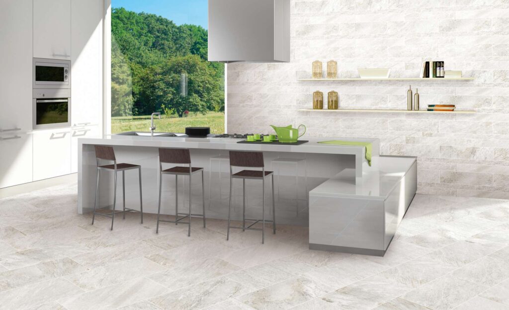Collezione Discovery Glacier nel formato 30x60: pavimento e rivestimento coordinati in gres porcellanato effetto pietra chiara per una cucina moderna e luminosa. La posa sfalsata delle piastrelle a pavimento e il rivestimento a muretto sulla parete di fondo creano un design geometrico e armonioso. Al centro della stanza spicca un'elegante isola cucina bianca lucida con sgabelli di design, affiancata da un'ampia vetrata che inonda lo spazio di luce naturale e offre una rilassante vista su un bosco verdeggiante.