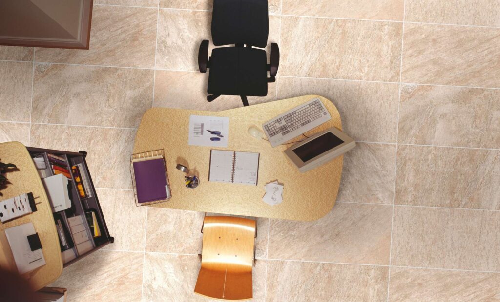 La collezione Discovery Sunwood, nel formato 30x60, riveste il pavimento di un elegante ambiente living adibito a spazio di lavoro o home office, catturato in una suggestiva vista dall'alto. Le piastrelle in gres porcellanato beige dalle calde sfumature e venature naturali sono posate in modo regolare, definendo la superficie su cui poggiano una scrivania da ufficio attrezzata con computer, sedia ergonomica nera e una sedia in legno, per un'atmosfera calda, luminosa e professionale.