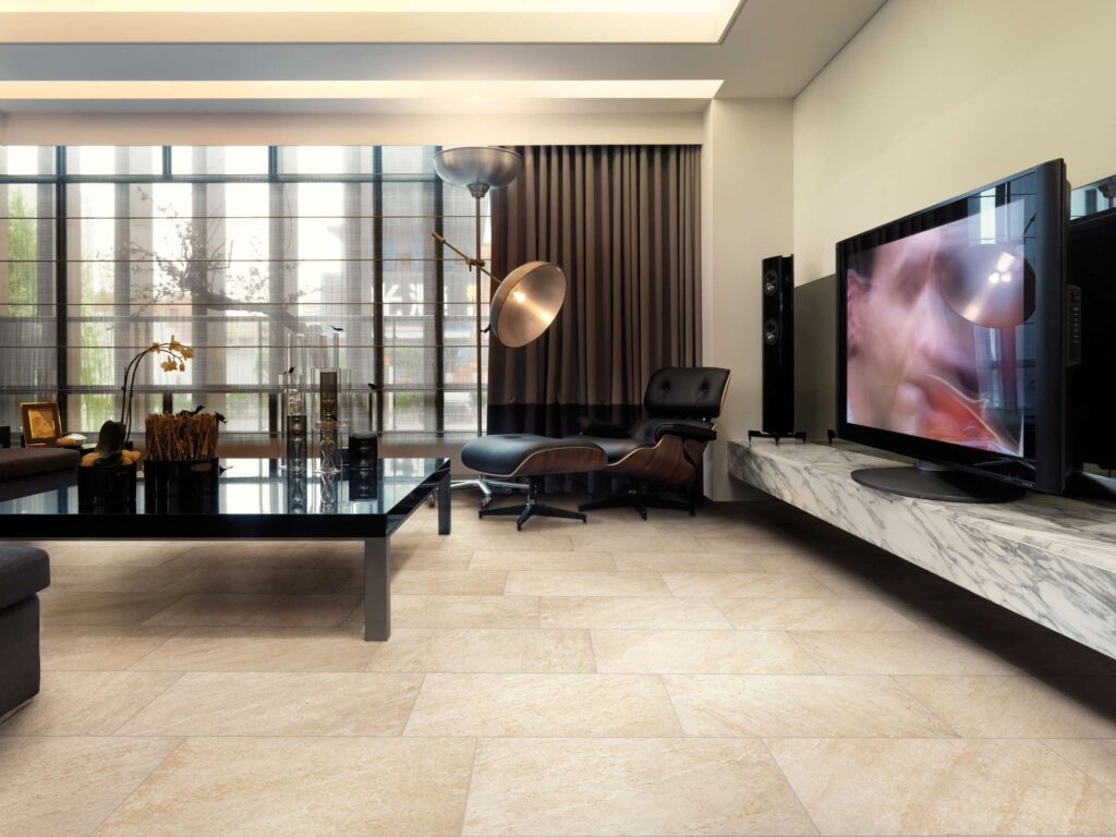 La collezione Discovery Sunwood nel formato 30x60 cm riveste il pavimento di un raffinato e moderno soggiorno con piastrelle effetto pietra naturale nei toni caldi del beige e del sabbia, posate con uno schema sfalsato a correre. L'elegante zona giorno è caratterizzata da arredi di design, tra cui una poltrona con poggiapiedi in pelle nera, un grande tavolo basso scuro e una consolle sospesa in marmo bianco sotto il televisore, creando un'atmosfera lussuosa e accogliente illuminata da ampie vetrate.