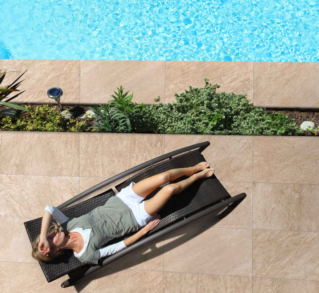Pavimentazione per esterni della collezione DISCOVERY SUNWOOD 30x60, caratterizzata da piastrelle in gres porcellanato effetto pietra nelle calde tonalità del sabbia. La vista dall'alto cattura un momento di relax a bordo piscina, con una donna distesa su un lettino prendisole in rattan scuro posizionato sulla pavimentazione. La posa regolare del formato 30x60 definisce con eleganza l'area solarium, arricchita da una rigogliosa aiuola decorativa che separa il patio dall'acqua azzurra e cristallina della piscina, evocando un'atmosfera estiva lussuosa e rigenerante.