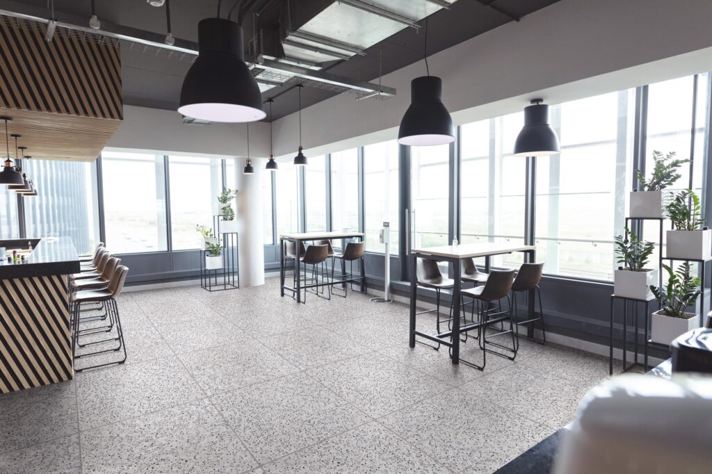 La collezione D_LASHh, nella variante Classic Shades in formato 90x90, veste il pavimento di un moderno e luminoso ristorante dal raffinato design contemporaneo. Il gres porcellanato effetto terrazzo, caratterizzato da una texture granulare grigia e posato in modo regolare, si integra armoniosamente in un ambiente industrial chic con ampie vetrate, tavoli alti, sgabelli scuri e grandi lampade a sospensione nere che valorizzano la luminosità dello spazio.