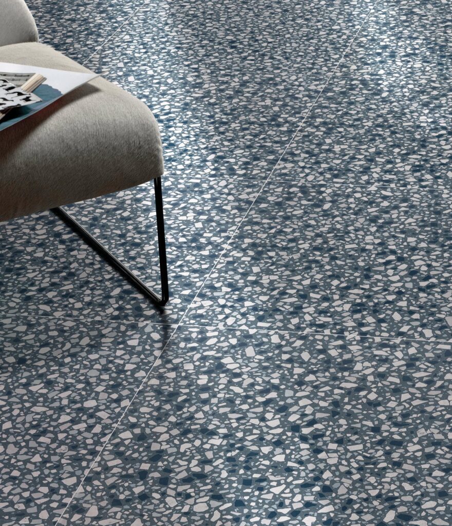La collezione D Lash presenta il gres porcellanato Mineral Blue nel formato 60x60, caratterizzato da un raffinato effetto terrazzo alla veneziana con graniglia bianca, azzurra e grigia su un fondo blu minerale. L'immagine mostra un dettaglio della pavimentazione posata con precisione, accostata a una moderna sedia imbottita in tessuto grigio chiaro con gambe in metallo nero, creando un'atmosfera contemporanea e luminosa grazie alla luce naturale che si riflette sulla superficie ceramica.