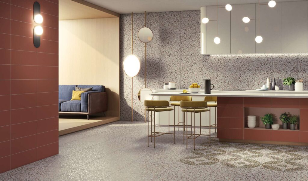La collezione D_Lash, nella variante colore Nordic Sand in formato 60x60 cm con elementi decorati (Dec.), caratterizza con eleganza questa cucina moderna e di design. Il pavimento e il paraschizzi a parete presentano piastrelle a effetto terrazzo veneziano nei toni chiari della sabbia, arricchiti sul pavimento da una posa decorativa geometrica a effetto tappeto. Al centro della stanza, l'isola cucina color terracotta con top bianco e sgabelli in velluto ocra si abbina armoniosamente alla parete laterale color ruggine. Sullo sfondo, un'apertura luminosa rivela un'accogliente zona giorno con divano blu.