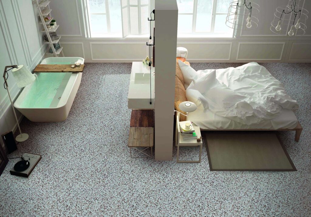La collezione D, nella sofisticata variante Lash Nordic Sand in formato 60x60 cm, riveste con eleganza il pavimento di un moderno e luminoso open space che unisce camera da letto e bagno di design. L'immagine mostra la posa continua di piastrelle in gres porcellanato effetto terrazzo veneziano, caratterizzate da una trama fine e multicolore su fondo chiaro. Lo spazio è diviso in modo funzionale da una parete centrale: a sinistra si apre la zona bagno con una raffinata vasca freestanding riempita d'acqua e un lavabo sospeso, mentre a destra si trova la zona notte con un letto matrimoniale dai tessuti bianchi. Grandi finestre sullo sfondo inondano l'ambiente di luce naturale, esaltando l'atmosfera nordica e minimale dell'ambiente.