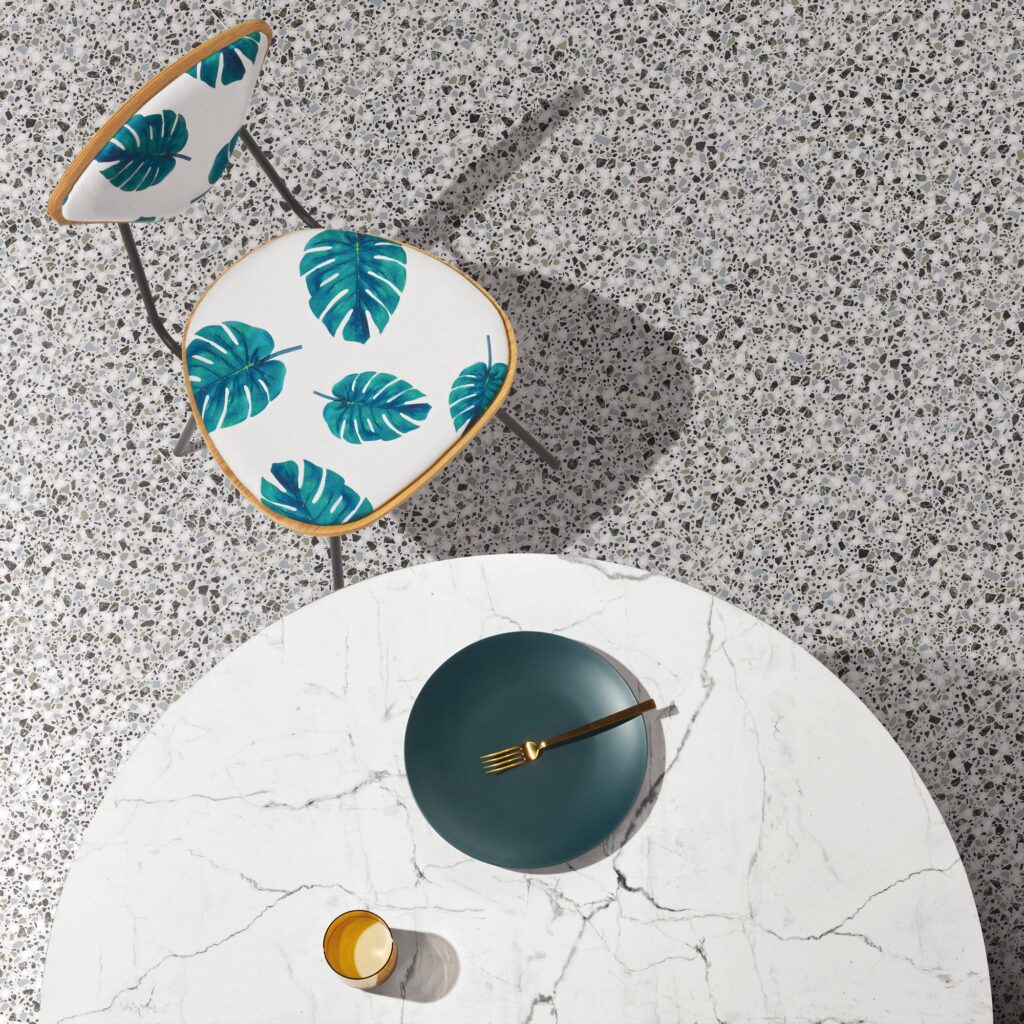 Dettaglio di pavimentazione effetto terrazzo per esterni Serie D_Lash colore Mountain Lake, con superficie in gres porcellanato dai toni grigio chiaro e inserti materici multiformi. Ambientazione contemporanea con tavolo effetto marmo e seduta decorata tropicale, ideale per spazi outdoor eleganti e moderni.