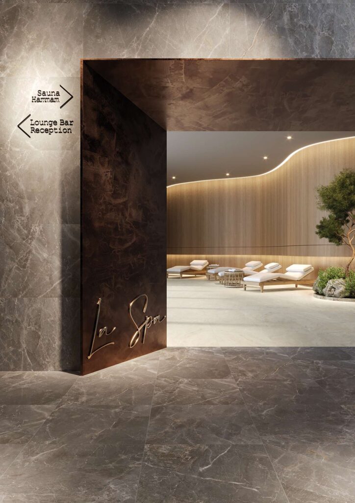 La collezione Elysion, proposta nei formati 60x60 e 60x120, caratterizza l'elegante ingresso di una spa d'eccellenza con pavimenti e rivestimenti in gres porcellanato effetto marmo grigio scuro, impreziositi da venature naturali e posati a regola d'arte. Un imponente portale in metallo scuro con la scritta dorata "La Spa" introduce a una luminosa zona relax interna, dove la pavimentazione è realizzata con la collezione Travertino nel formato 60x120 dall'effetto pietra beige calda. L'ambiente benessere è completato da raffinati lettini in legno con cuscini bianchi, pareti in legno retroilluminate e un'atmosfera lussuosa e rilassante.