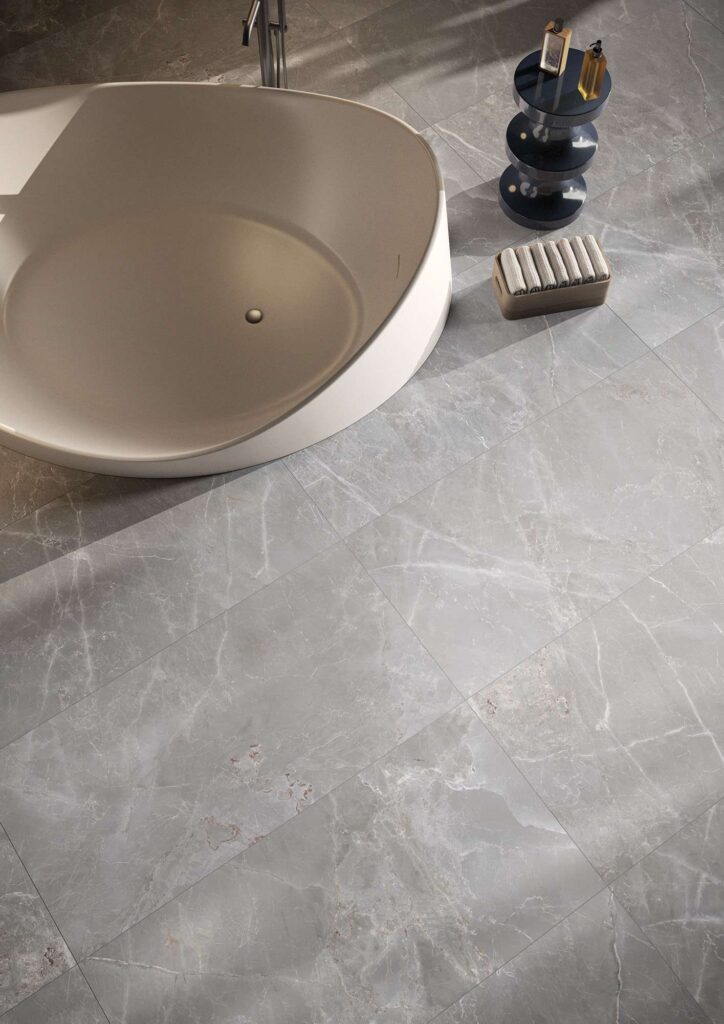 La collezione Elysion Grey nel formato 60x120 cm riveste con eleganza il pavimento di un bagno di design in questo scatto ravvicinato. Le piastrelle in gres porcellanato grigio, caratterizzate da raffinate venature calde ed effetto pietra naturale, incorniciano una moderna vasca freestanding circolare. Accanto alla vasca, la posa impeccabile è valorizzata da elementi d'arredo minimalisti, come un tavolino di design con flaconi e un piccolo contenitore in legno con asciugamani arrotolati, che evocano un'atmosfera di lussuoso benessere.