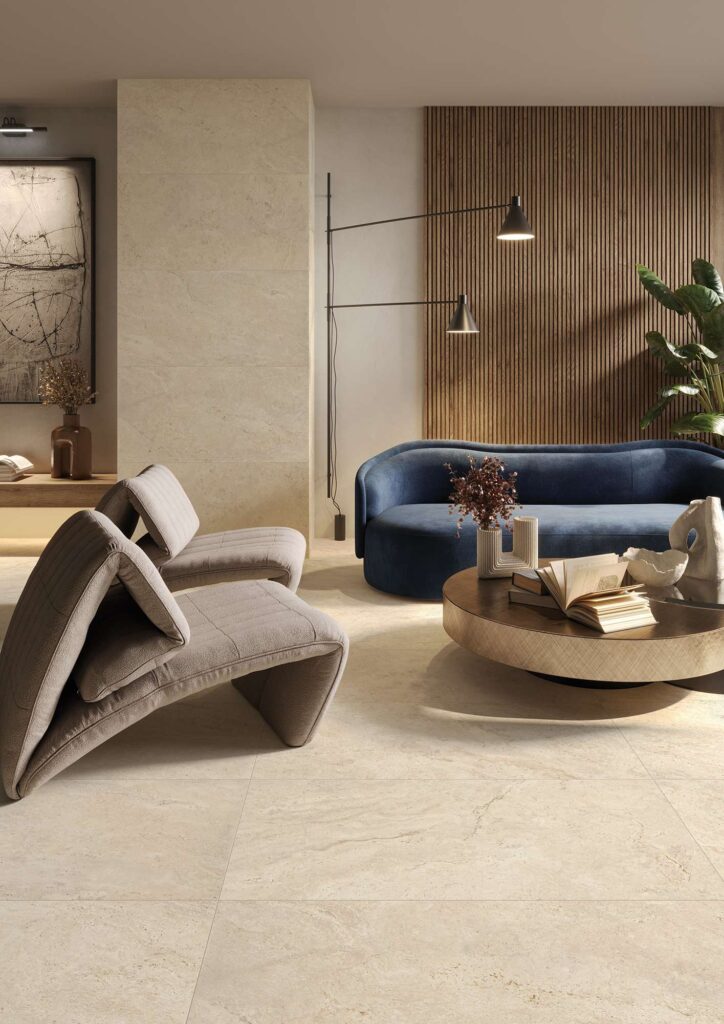 La collezione Elysion nel formato 60x120 veste con eleganza il pavimento e la parete di un luminoso ambiente living moderno. Le piastrelle in gres porcellanato effetto pietra travertino beige, dalle sfumature calde e naturali, creano una superficie continua e armoniosa. Lo spazio è arredato in stile contemporaneo: in primo piano spiccano due poltrone di design grigie, affiancate da un tavolino tondo in legno e da un divano blu notte accostato a una parete decorata con listelli di legno verticali, per un'atmosfera calda, lussuosa e accogliente.