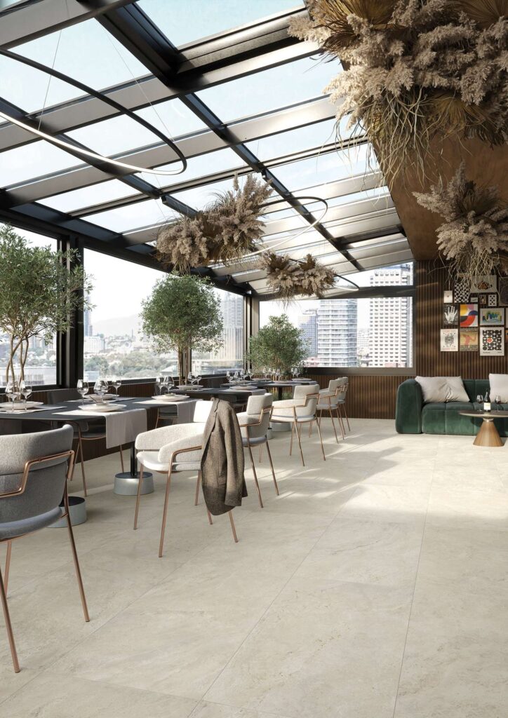 La collezione Elysion si esprime al meglio in questa ambientazione ristorante, dove il pavimento in gres porcellanato effetto travertino nel formato 60x120 cm dona luminosità e continuità visiva. Lo spazio, concepito come una raffinata veranda con soffitto in vetro e ampie vetrate sulla città, è arredato con tavoli moderni, eleganti sedie dal design contemporaneo e piante decorative di ulivo. La posa regolare delle grandi lastre ceramiche nei toni caldi del beige chiaro esalta l'atmosfera sofisticata, ariosa e naturale del locale.