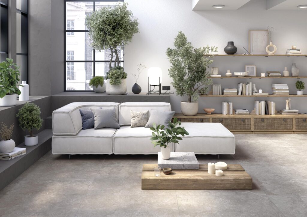 Collezione G nella variante Stone Dorset Grey: pavimento in gres porcellanato effetto pietra che riveste con eleganza un ampio e luminoso soggiorno moderno. L'immagine mette in risalto la posa di grandi formati quadrati dalle sfumature grigie naturali e materiche, che donano continuità visiva all'ambiente. L'arredo è composto da un divano modulare bianco, mensole in legno naturale ricche di libri e oggetti di design, e numerose piante ornamentali che infondono un senso di benessere e freschezza, completando un'atmosfera ricercata e contemporanea.