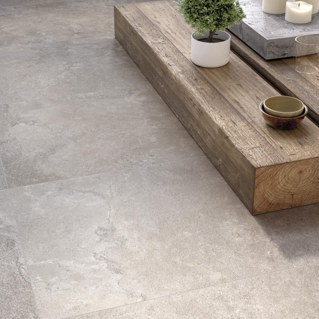 Collezione G, variante Dorset Grey: pavimento in gres porcellanato effetto pietra in grandi formati che riveste con eleganza un ambiente living moderno. La posa lineare delle piastrelle grigie, caratterizzate da una texture naturale e materica, è accostata a elementi d'arredo in legno grezzo e decorazioni minimali come una piccola pianta e candele, creando un'atmosfera accogliente e sofisticata.