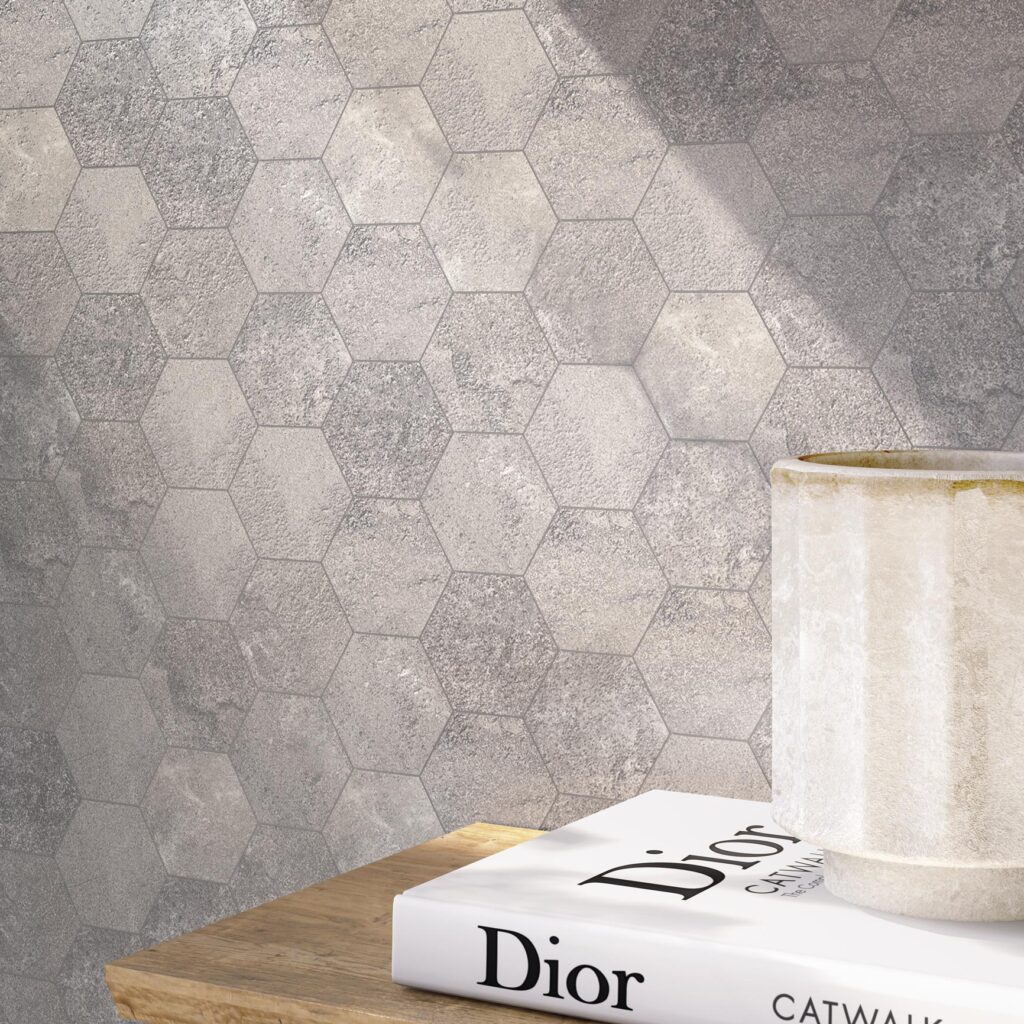 Collezione G, piastrelle effetto pietra nella variante colore Dorset Grey, qui proposte in un elegante formato esagonale. L'immagine mostra un raffinato dettaglio d'interni in cui la parete, rivestita con questo mosaico esagonale dalle sfumature grigie e materiche, fa da sfondo a un piano in legno su cui poggiano un libro di Dior e un vaso in ceramica chiara, il tutto avvolto da una calda luce naturale.
