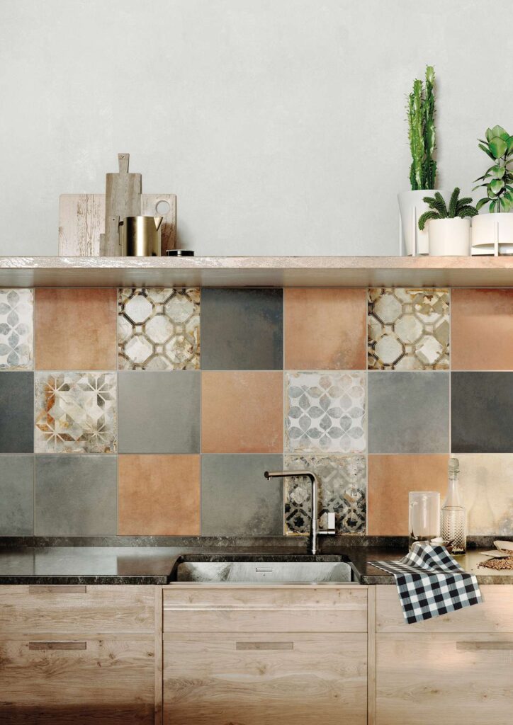 La collezione Heartland colore Amber 30x30, completa di raffinati decori, valorizza la parete di questa cucina con un backsplash dal fascino caldo e contemporaneo. Le piastrelle in gres porcellanato nel formato 30x30 cm sono posate in una composizione che alterna tonalità calde color ambra e terracotta a sfumature grigie, arricchite da elementi decorati con motivi geometrici e floreali dal gusto vintage. La parete piastrellata fa da sfondo a un moderno lavello incassato in un top scuro, coordinandosi armoniosamente con le basi in legno chiaro e una mensola superiore decorata con piante e accessori da cucina.