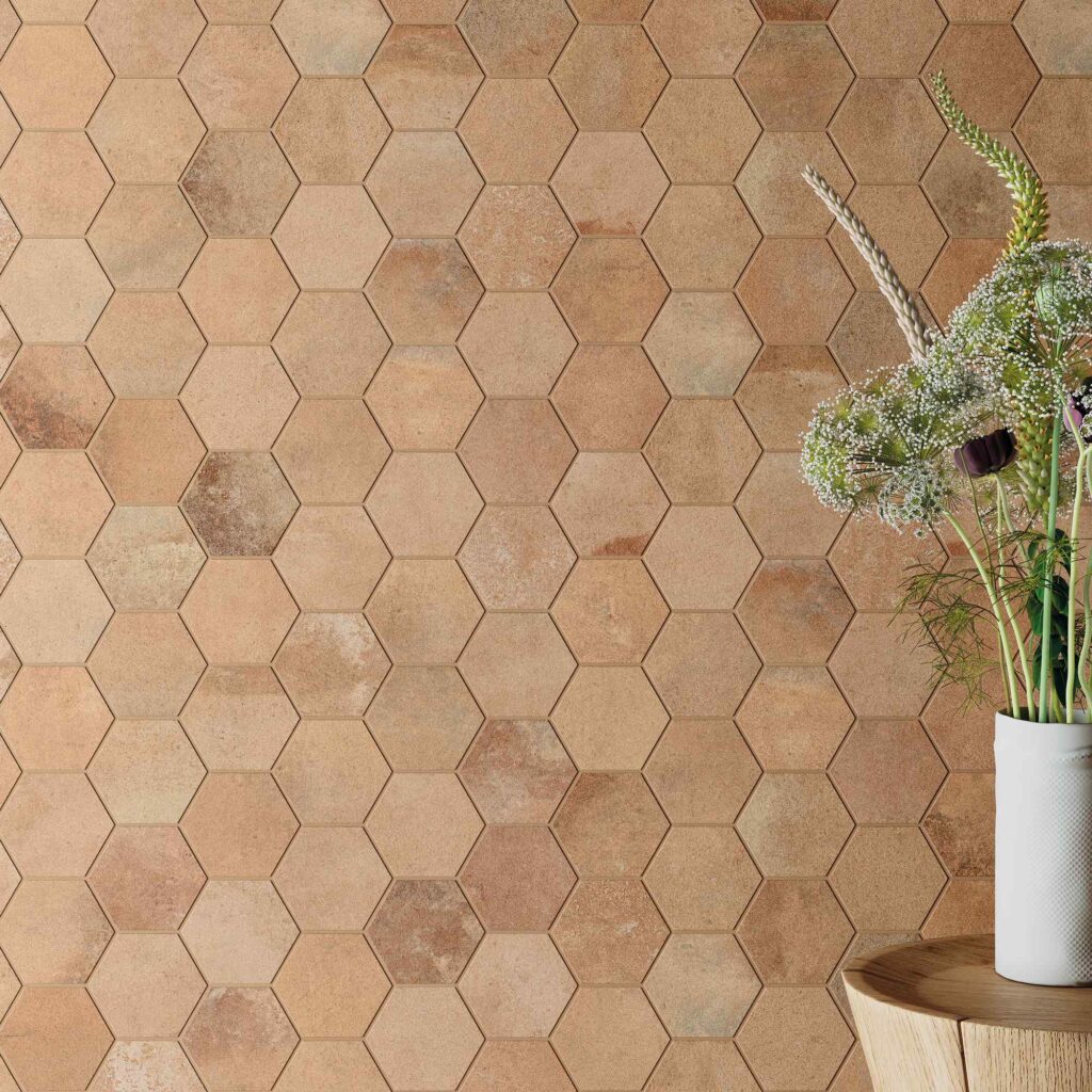 La collezione Heartland colore Amber mostra un primo piano del suo mosaico esagonale (Mos.Esag.) per rivestimenti, caratterizzato da tessere dalle calde sfumature color ambra, sabbia e terracotta dall'effetto materico e naturale. Sulla destra, la composizione è arricchita da un elegante vaso bianco con fiori di campo posato su uno sgabello in legno chiaro, che enfatizza l'atmosfera calda, accogliente e dal design organico dell'ambiente.