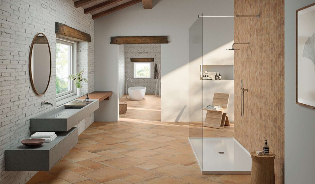 La collezione HEARTLAND colore AMBER in gres porcellanato effetto cotto arreda un ampio bagno luminoso dal design rustico contemporaneo. Il pavimento è posato in un suggestivo schema multiformato diagonale che valorizza le sfumature calde color ambra, mentre la parete interna della moderna doccia walk-in è rivestita con il mosaico esagonale coordinato. L'ambiente, caratterizzato da soffitti con travi in legno e una parete in mattoncini bianchi con lavabo sospeso in cemento, si apre sullo sfondo verso una zona benessere con vasca freestanding, evocando un'atmosfera accogliente, naturale e di assoluta eleganza.