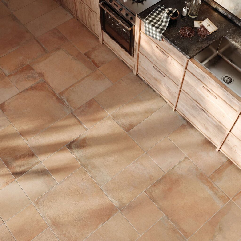 La collezione Heartland colore Amber Multiformato riveste il pavimento di una moderna cucina rustica con piastrelle in gres porcellanato effetto cotto dalle calde sfumature ambrate. La vista dall'alto mette in risalto la posa modulare multiformato, esaltata da una calda luce solare radente che disegna ombre geometriche sulla superficie. Accanto al pavimento, si inserisce una cucina lineare con basi in legno chiaro, un top scuro con lavello integrato e un forno da incasso, creando un'atmosfera accogliente e dal design d'eccellenza.