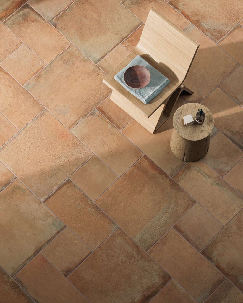 La collezione Heartland colore Amber in posa multiformato riveste il pavimento con calde piastrelle in gres porcellanato effetto cotto, caratterizzate da sfumature ambrate, aranciate e ruggine che donano un senso di calore e naturalezza. La vista dall'alto mette in risalto il raffinato schema di posa modulare a diverse dimensioni, sul quale poggiano una sedia di design in legno chiaro con un asciugamano e una ciotola, affiancata da un tavolino a tronco d'albero con accessori per il benessere. L'illuminazione morbida crea un'atmosfera rilassante, elegante e accogliente.