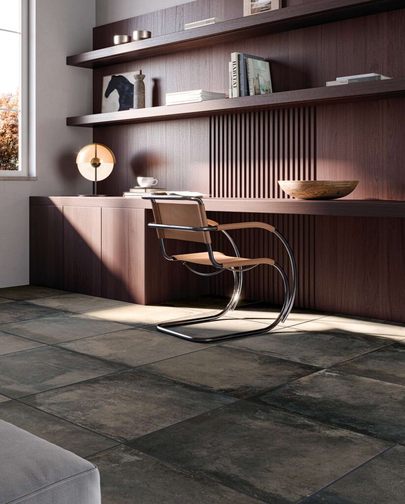La collezione Heartland, nella tonalità Charcoal e formato 60,3x60,3, riveste il pavimento di un raffinato studio domestico. Le piastrelle in gres porcellanato scuro, caratterizzate da una texture materica e sfumature naturali, sono posate in modo ortogonale e colpite da un fascio di luce solare radente. L'ambiente, dall'atmosfera calda e professionale, presenta una parete attrezzata in legno scuro con scrivania integrata, mensole decorate e un'iconica sedia di design in metallo cromato e cuoio.