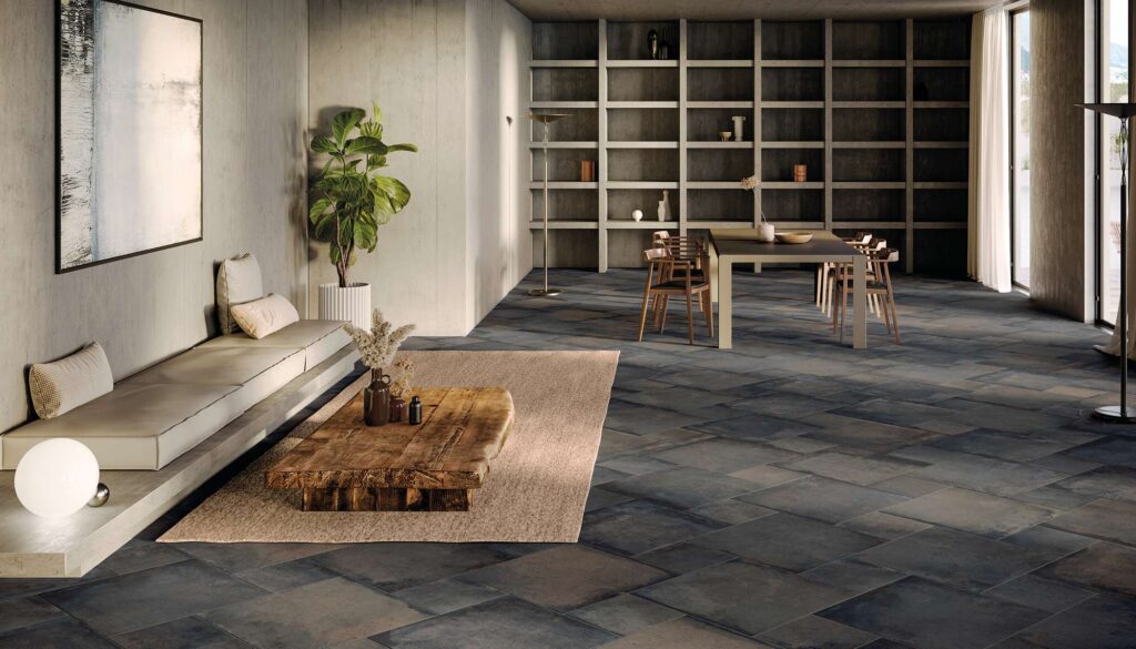 La collezione Heartland, qui nella variante cromatica Charcoal, veste il pavimento in gres porcellanato di questo raffinato ambiente living attraverso una dinamica posa multiformato. Lo scatto cattura un ampio e luminoso open space dal design contemporaneo e minimalista: le piastrelle nei toni profondi del grigio scuro effetto pietra si abbinano armoniosamente a pareti in cemento a vista, su cui risaltano una panca lineare con seduta chiara, un tavolino in legno massiccio grezzo e una pianta da interni. Sullo sfondo, la zona pranzo è elegantemente arredata con tavolo e sedie di design davanti a un'ampia libreria integrata a tutta parete, inondata dalla luce naturale che filtra dalle grandi vetrate.