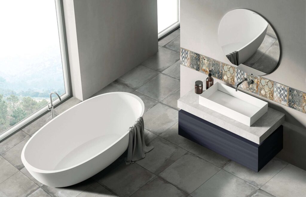 La collezione Heartland colore Clay, con piastrelle in gres porcellanato nel formato 60,3x60,3, riveste il pavimento di questo moderno e luminoso bagno di design. L'ambiente, caratterizzato da un'atmosfera minimale e sofisticata, ospita una vasca freestanding bianca di forma ovale posizionata accanto a un'ampia vetrata, e un mobile lavabo sospeso con specchio circolare. Sulla parete di fondo spicca una raffinata fascia ornamentale realizzata con i decori nel formato 30x30, caratterizzati da eleganti trame geometriche e tonalità calde che donano colore e personalità alla stanza.