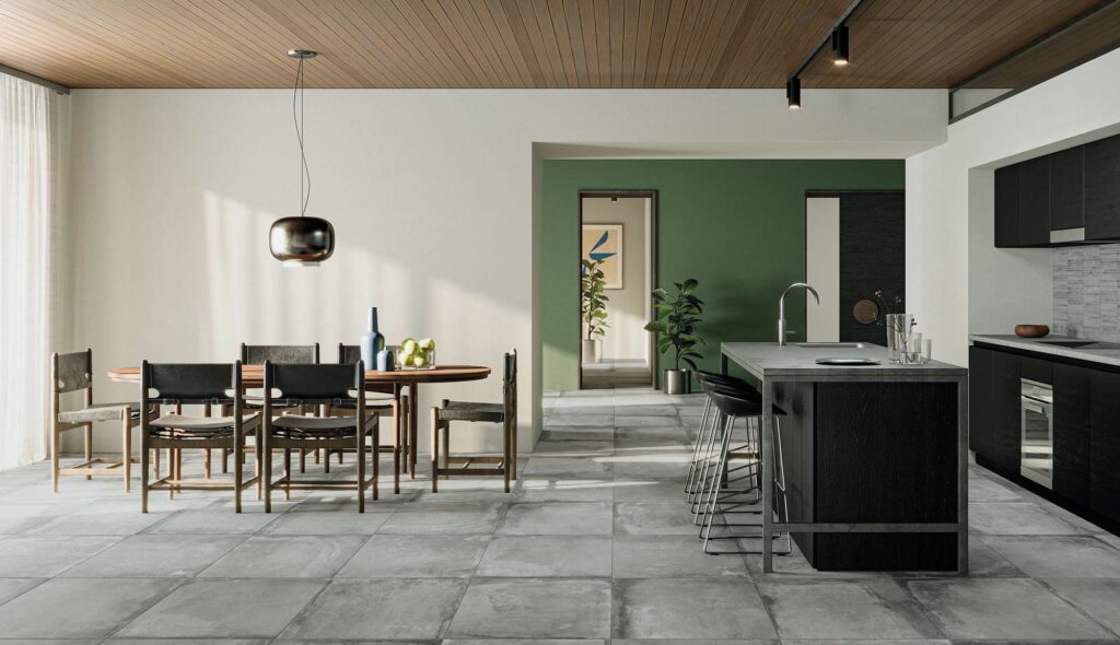 La collezione Heartland colroe Clay, qui presentata nel formato 60,3x60,3 cm, riveste con eleganza il pavimento di un moderno e luminoso open space con cucina e zona pranzo. Le grandi piastrelle in gres porcellanato nella calda tonalità argilla valorizzano l'ambiente, caratterizzato da un soffitto con doghe in legno, un'isola cucina nera e una parete di fondo verde salvia. Sulla destra, la zona cottura è completata da un paraschiena decorato con il mosaico a listelli MOS.STICKS, che crea un raffinato contrasto materico con la posa regolare e minimalista del pavimento.