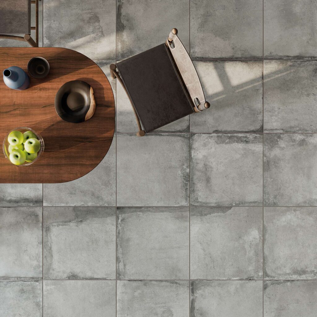 Collezione Heartland Clay nel formato 60,3x60,3 cm: pavimento in gres porcellanato dall'aspetto materico e naturale, ideale per un ambiente cucina raffinato. La vista dall'alto mette in risalto la posa geometrica delle piastrelle nei toni del grigio e dell'argilla con bordi ed effetti stonalizzati. Completano la scena un tavolo in legno scuro arricchito da una ciotola di mele verdi e stoviglie di design, e una sedia in cuoio scuro, il tutto avvolto da una calda luce naturale che proietta ombre delicate sul pavimento.