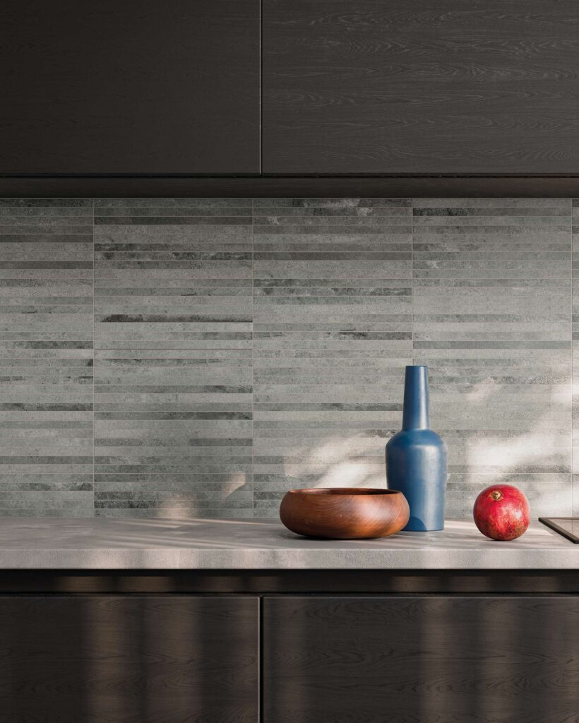 La collezione HEARTLAND colore CLAY nel formato mosaico a listelli (MOS.STICKS) riveste la parete paraschizzi di una cucina moderna e raffinata. Le piastrelle in gres porcellanato presentano calde sfumature di grigio ed effetto pietra naturale con posa orizzontale. Sul top cucina grigio chiaro, illuminato da una morbida luce solare filtrante, sono adagiati un vaso blu decorativo, una ciotola in legno e una melagrana, il tutto incorniciato da eleganti mobili in legno scuro.