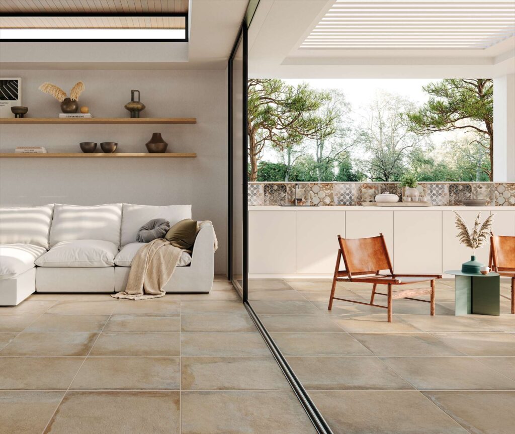 La collezione Heartland colore Sand nel formato 60,3x60,3 cm veste con continuità ed eleganza un luminoso ambiente indoor-outdoor. L'immagine mostra una raffinata zona giorno con un divano bianco e mensole in legno, che si apre in modo fluido verso un patio esterno attraverso una grande vetrata scorrevole. Il pavimento in gres porcellanato color sabbia, posato in linea, si estende senza interruzioni sul terrazzo, dove risaltano sedute di design in legno e un backsplash con piastrelle decorate per la cucina esterna, creando un'atmosfera calda, naturale e profondamente contemporanea.