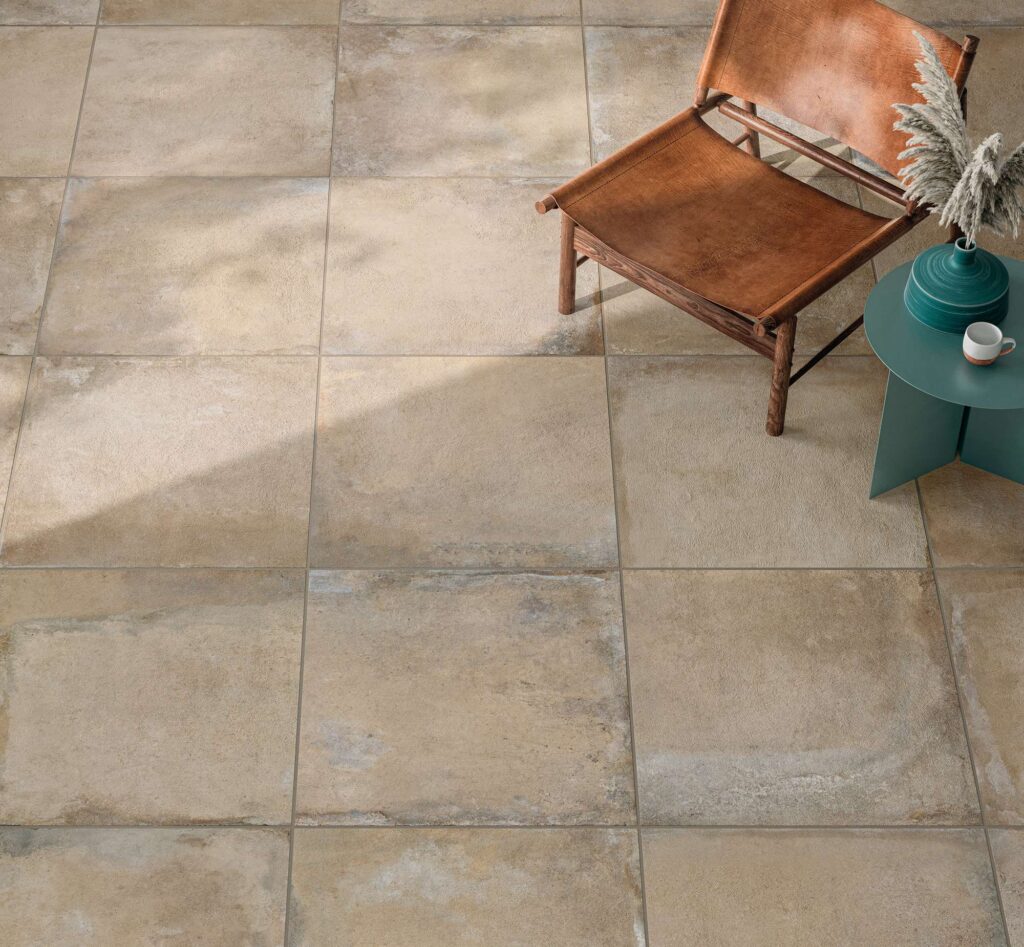 La collezione Heartland colore Sand nel formato 60,3x60,3 cm valorizza questo spazio interno con un pavimento in gres porcellanato effetto pietra dalle calde tonalità del sabbia e del beige. L'inquadratura ravvicinata mette in mostra la ricca texture materica e le stonalizzazioni naturali delle piastrelle posate a regola d'arte. L'atmosfera accogliente è completata da una poltrona di design in legno e cuoio marrone e da un tavolino circolare verde petrolio con elementi decorativi, il tutto impreziosito da un gioco di luci e ombre naturali.