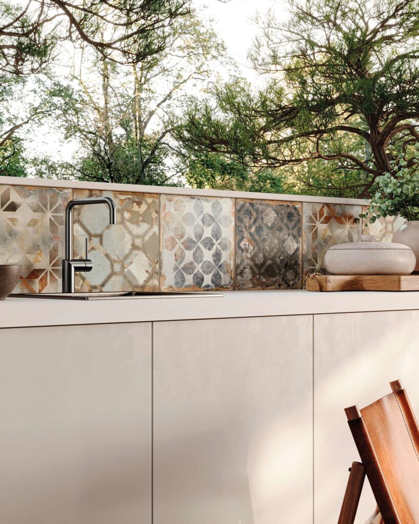 La collezione HEARTLAND colore SAND DECORI veste l'alzatina di una raffinata cucina da esterno in un patio, con piastrelle in formato quadrato caratterizzate da suggestivi motivi geometrici e floreali in stile cementine. Le sfumature calde del sabbia, del beige e del ruggine si alternano a toni più freddi del grigio e dell'antracite dall'effetto piacevolmente vissuto, creando un contrasto armonioso con il moderno top chiaro e il miscelatore scuro di design. Sullo sfondo, la luce naturale del sole filtra tra la vegetazione, esaltando l'atmosfera elegante e rilassante dell'ambiente outdoor.