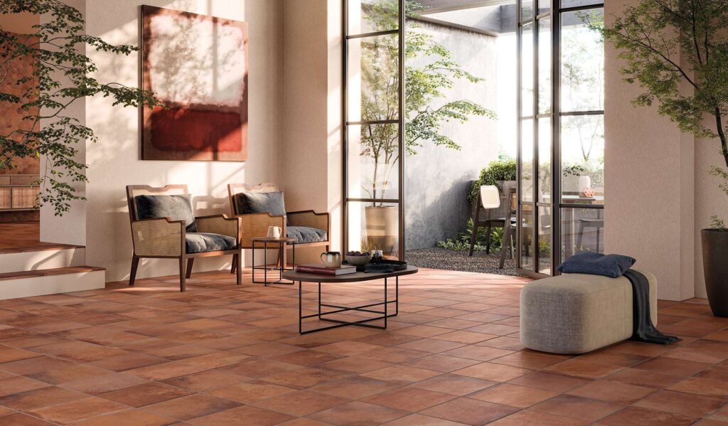 La collezione Heartland Terracotta nel formato 30x30 valorizza il pavimento di questo elegante e luminoso soggiorno (living). Le piastrelle in gres porcellanato effetto cotto, caratterizzate da calde sfumature naturali, sono posate a giunto unito e si estendono in un ambiente accogliente arredato con poltrone in legno e paglia di Vienna, un tavolino da caffè e un morbido pouf. Grandi vetrate aperte su un patio esterno inondano la stanza di luce solare, creando un'armoniosa continuità tra interno ed esterno.