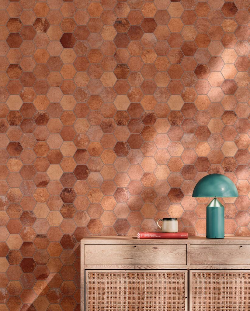 La collezione HEARTLAND TERRACOTTA nel formato mosaico esagonale riveste con eleganza una parete interna, esaltando le calde sfumature della terra e del cotto tradizionale in chiave contemporanea. L'ambientazione mostra un raffinato living arricchito da una madia in legno chiaro con dettagli in paglia di Vienna, su cui poggiano una lampada di design verde petrolio, un libro e una tazza in ceramica. La luce naturale filtra creando delicate ombre sulla superficie ceramica, regalando un'atmosfera accogliente, intima e di grande pregio estetico.
