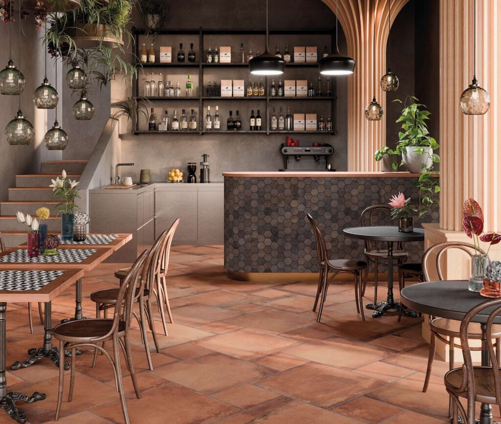 La collezione Heartland Terracotta, posata in un caldo e dinamico schema multiformato, caratterizza il pavimento di un accogliente e raffinato ristorante dal design contemporaneo. Lo spazio è valorizzato dal bancone bar curvo, impreziosito dal rivestimento in mosaico esagonale nella variante Charcoal, che crea un elegante contrasto materico e cromatico. L'atmosfera in stile bistrot è completata da tavoli neri, sedie in legno scuro, rigogliose piante decorative e un'illuminazione soffusa che esalta le sfumature naturali e la texture delle superfici ceramiche.