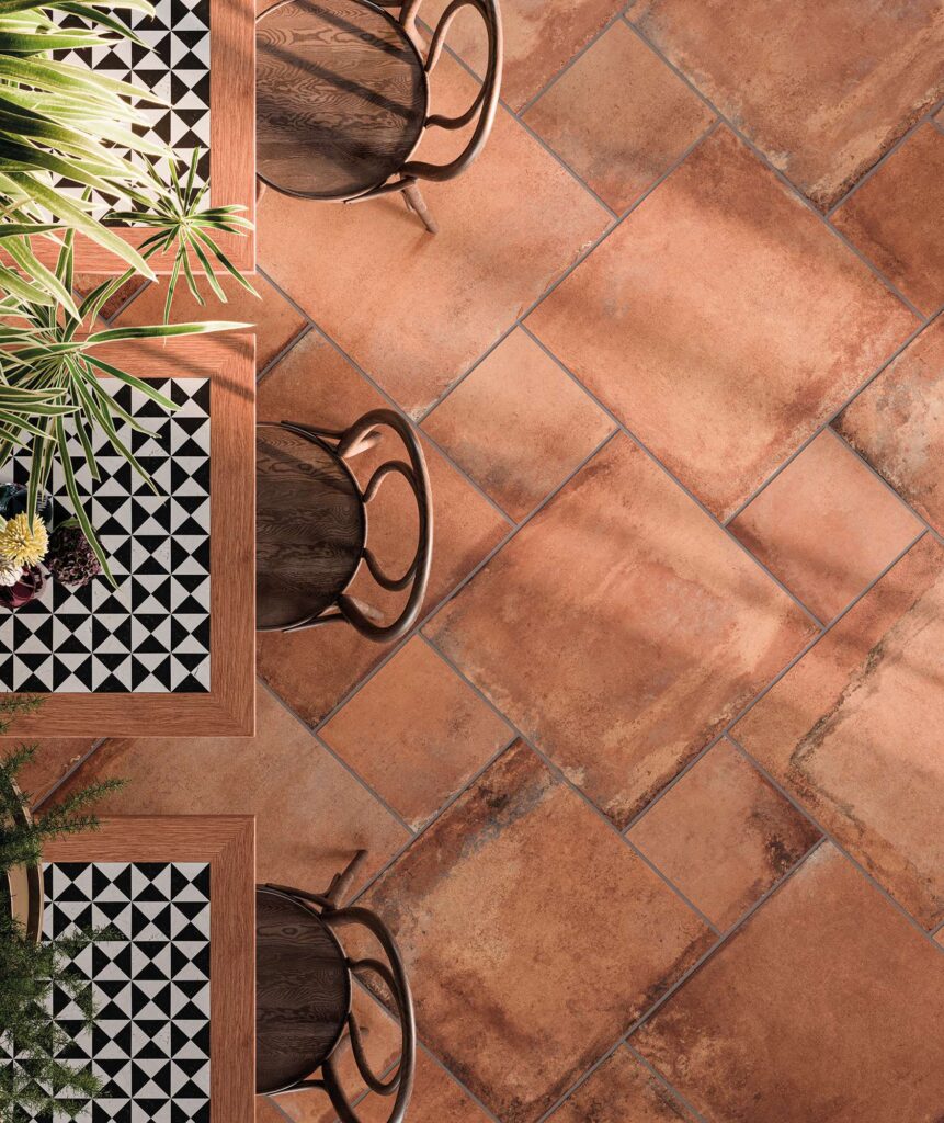 La collezione HEARTLAND in posa multiformato definisce lo stile di questo raffinato ristorante, ritratto in una suggestiva veduta dall'alto. Le piastrelle in gres porcellanato colore TERRACOTTA effetto cotto, caratterizzate da calde sfumature terrose e stonalizzazioni naturali, sono posate secondo uno schema modulare dinamico. Sulla sinistra, eleganti tavoli in legno con top dai pattern geometrici bianchi e neri si accostano a sedie curve in legno scuro e piante verdi rigogliose, creando un'atmosfera calda, accogliente e di grande design.