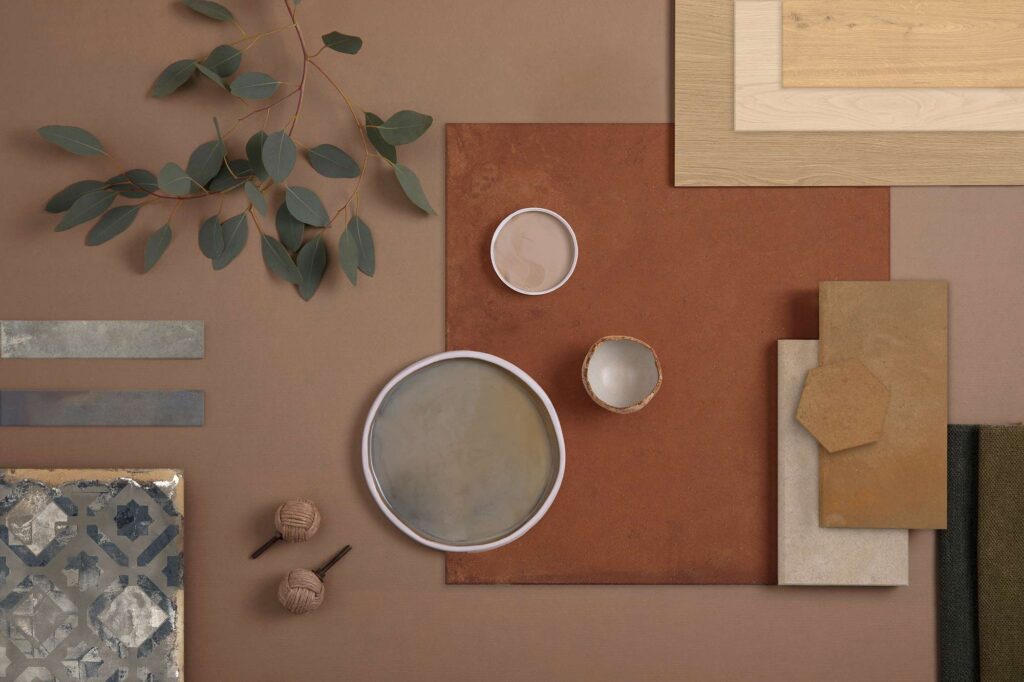 La collezione HEARTLAND è protagonista di un raffinato moodboard ispirazionale in stile flat lay, dominato dalle calde tonalità della terra come il terracotta, l'ocra, il sabbia e dettagli grigio-azzurri. La composizione mette in mostra la ricca varietà di formati e superfici, tra cui piastrelle quadrate di grandi dimensioni, listelli rettangolari, elementi esagonali, campioni effetto legno e una formella ceramica decorata con motivi geometrici dal sapore artigianale. Accanto ai campioni ceramici, elementi naturali e di design completano la scena: un ramo di eucalipto dalle foglie verdi, ciotole in ceramica, pomelli in corda intrecciata e tessuti materici nei toni del verde oliva, evocando un'atmosfera elegante, calda e contemporanea per l'interior design.