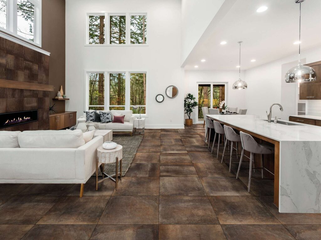 La collezione Hemisphere Copper 60x60 valorizza con eleganza questo ampio e luminoso living open space dal design contemporaneo. Il pavimento in gres porcellanato effetto metallo presenta calde sfumature brunite color rame (copper), posate in modo regolare per unire armoniosamente l'area relax, arredata con raffinati divani bianchi e un moderno camino a parete, e la zona cucina, caratterizzata da una grande isola in marmo bianco con sgabelli di design. Grandi vetrate sullo sfondo inondano lo spazio di luce naturale, esaltando la texture materica e la superficie riflettente delle piastrelle.