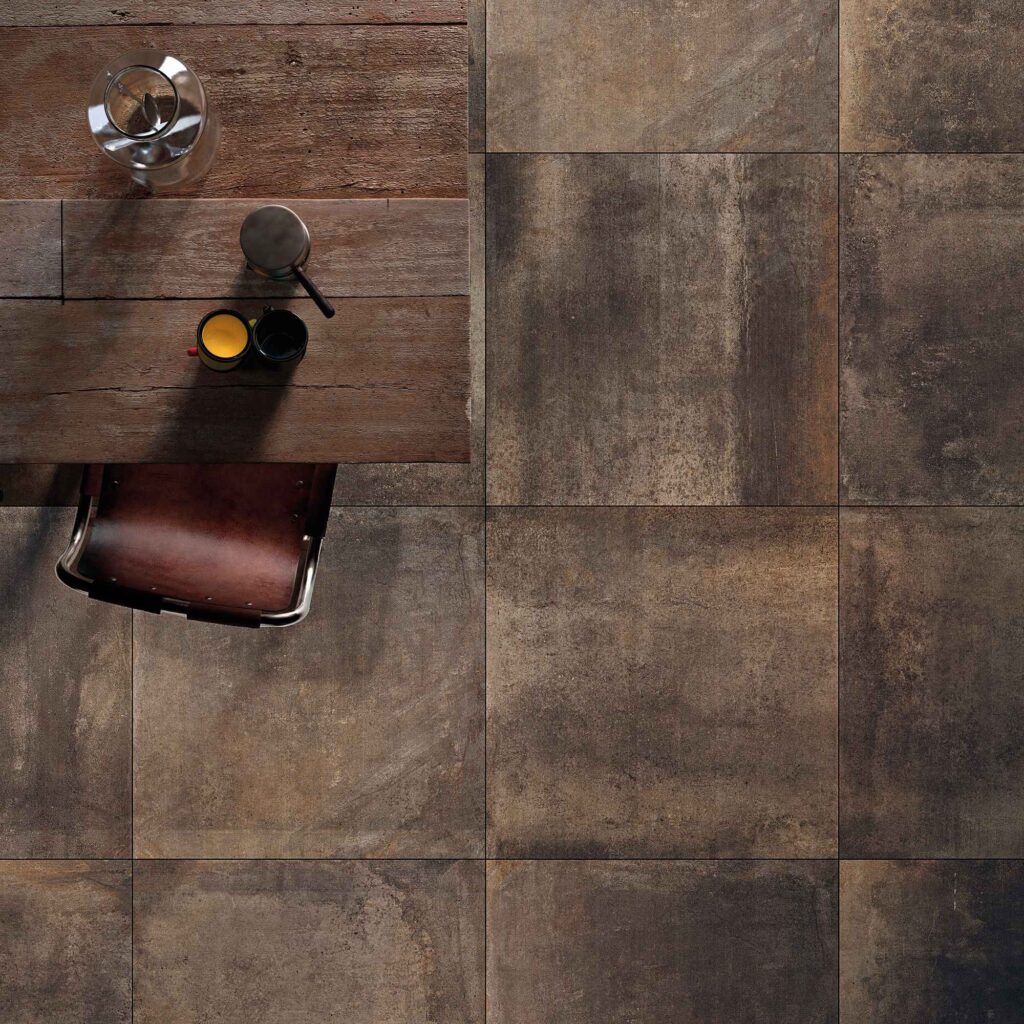 La collezione Hemisphere, nella calda variante cromatica Copper e formato 80x80 cm, è protagonista di questa raffinata vista dall'alto che esalta il design del gres porcellanato effetto metallo ossidato. Le piastrelle, caratterizzate da profonde sfumature ramate, brune e scure, sono posate in modo lineare e accostate a un tavolo in legno grezzo e a una sedia in cuoio marrone, ricreando un'atmosfera d'interni elegante, accogliente e dal forte carattere industrial chic.