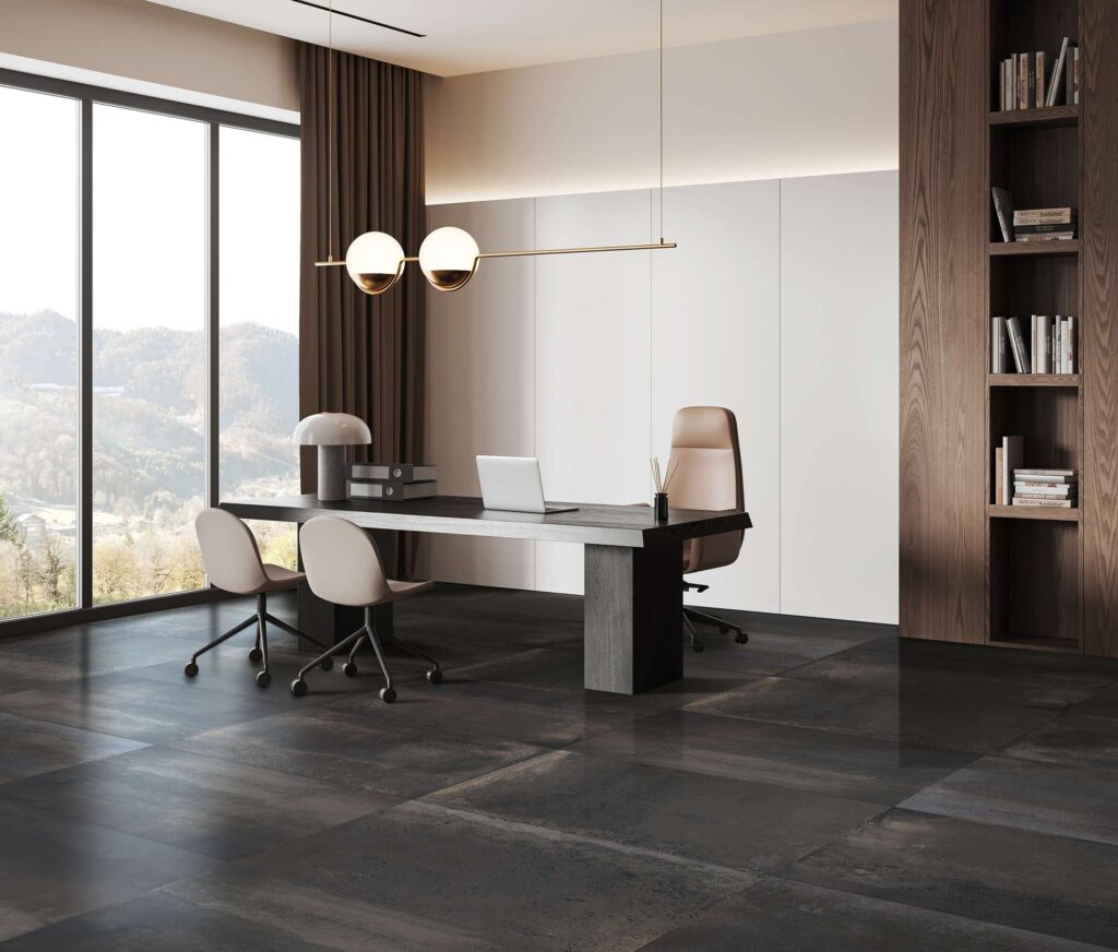 La collezione Hemisphere nella variante Iron, nel formato 60x120 cm, caratterizza il pavimento di questo ufficio moderno e raffinato attraverso lastre in gres porcellanato scuro dall'elegante effetto metallico. L'ambiente, luminoso e sofisticato, è arredato con una scrivania direzionale in legno scuro, sedie di design nei toni del beige, una libreria a muro e un lampadario minimale con dettagli dorati. Sullo sfondo, una grande vetrata a tutta altezza rivela una suggestiva vista panoramica sulle colline, valorizzando i toni profondi e la texture materica della pavimentazione.