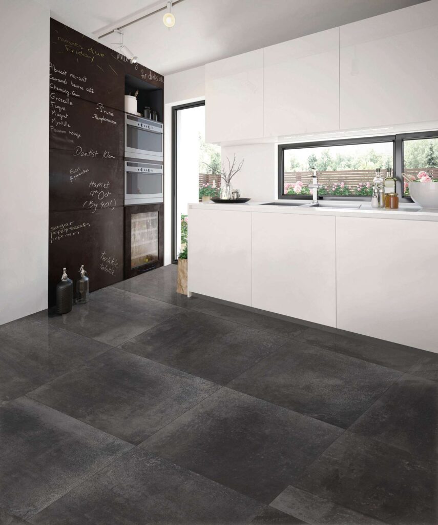 La collezione HEMISPHERE, nella variante colore Iron e formato 80x80 cm, caratterizza il pavimento di una cucina moderna e luminosa. Le grandi piastrelle scure effetto metallo, posate in modo regolare, creano un elegante contrasto cromatico con i mobili minimalisti laccati in bianco lucido e la parete scura stile lavagna attrezzata con elettrodomestici. L'ambiente è inondato di luce naturale proveniente dalle ampie vetrate aperte sul giardino, esaltando la texture materica e la raffinatezza contemporanea della superficie ceramica.