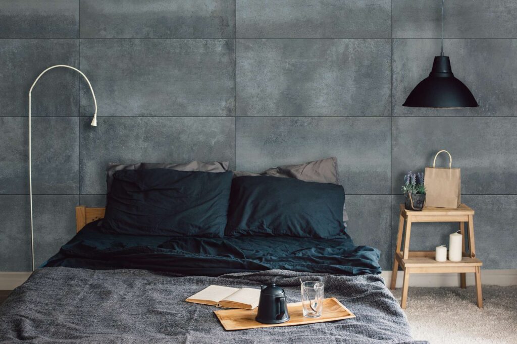 La collezione Hemisphere Lagune nel formato 60x120 cm riveste la parete di questa raffinata camera da letto, mostrando grandi lastre in gres porcellanato dall'effetto materico e dalle profonde sfumature grigio-bluastre, posate in senso orizzontale. L'ambiente, dall'atmosfera intima e moderna, presenta un letto coordinato con lenzuola scure, arricchito da un vassoio in legno con teiera e libro in primo piano. Ai lati, una minimale lampada ad arco bianca sulla sinistra e un comodino in legno con candele e una lampada a sospensione nera sulla destra completano il design contemporaneo e sofisticato.