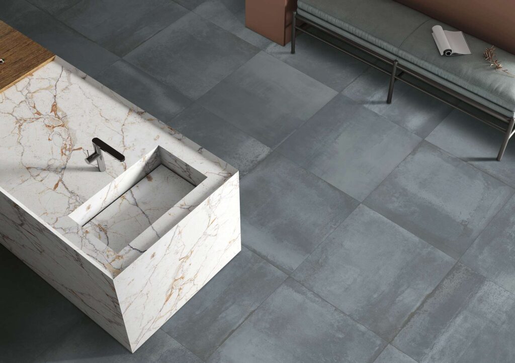 La collezione Hemisphere Lagune, nel formato 60x60 cm, riveste il pavimento di un'elegante cucina moderna con piastrelle in gres porcellanato dalle tonalità grigio-azzurre intense ed effetto materico. La vista dall'alto mette in mostra la posa regolare e l'accostamento armonioso con un'isola cucina di design in gres effetto marmo bianco dalle calde venature dorate, completa di lavello integrato e miscelatore nero. Sullo sfondo, una panca minimalista e un dettaglio a parete color terracotta completano l'atmosfera sofisticata e contemporanea dell'ambiente.