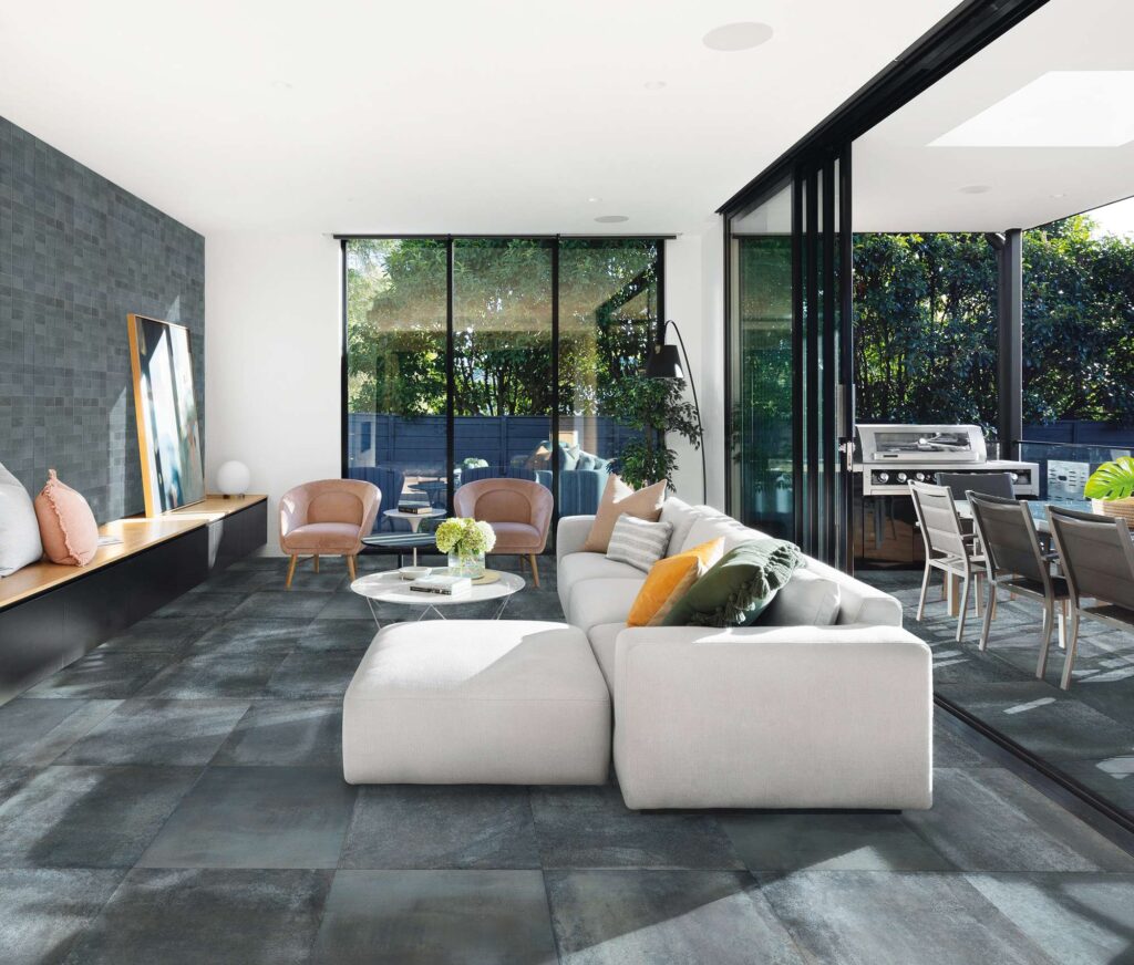 La collezione Hemisphere Lagune nel formato 60x60 definisce il pavimento di questo moderno e luminoso soggiorno con piastrelle in gres porcellanato dalle intense sfumature scure e metalliche, posate in modo regolare. L'ambiente living, caratterizzato da ampie vetrate che si aprono su un patio esterno, è arredato con un divano modulare chiaro, poltroncine rosa antico e un tavolino da caffè. Sulla parete di sinistra, un elegante rivestimento a mosaico scuro crea un raffinato contrasto geometrico.