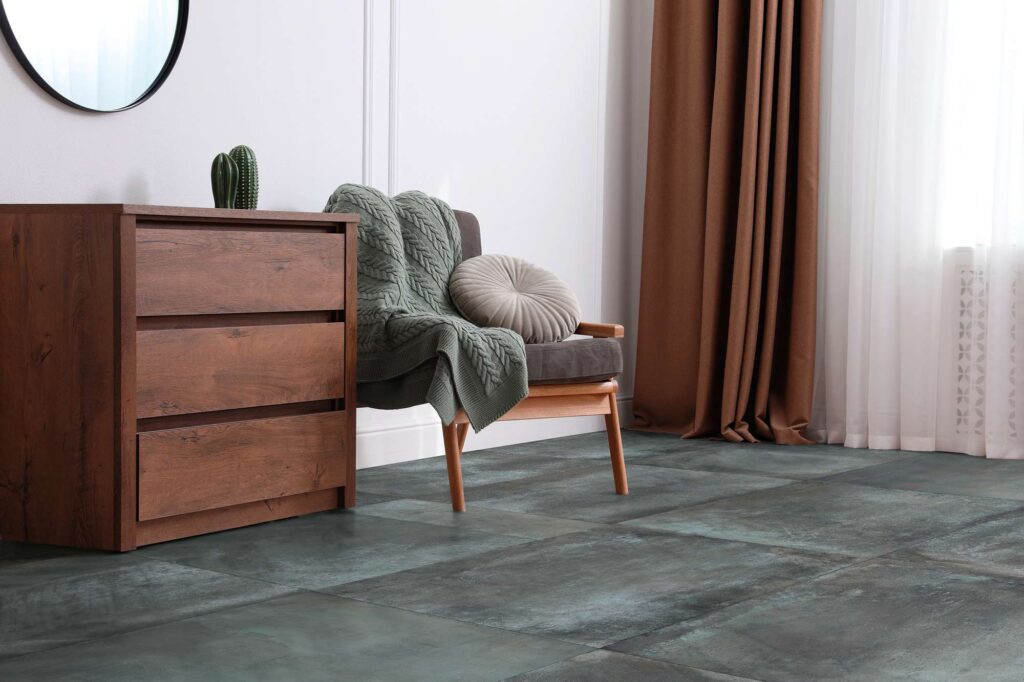 La collezione HEMISPHERE, nella raffinata variante colore LAGUNE e formato 80X80 cm, definisce lo stile di questo elegante soggiorno contemporaneo. La pavimentazione in gres porcellanato dalle profonde sfumature verde laguna metallizzato è posata in modo lineare, offrendo un sofisticato contrasto cromatico con gli arredi caldi: una cassettiera in legno scuro, una poltrona impreziosita da una morbida coperta lavorata a maglia verde salvia e ampie tende color terra, per un'atmosfera intima e di classe.