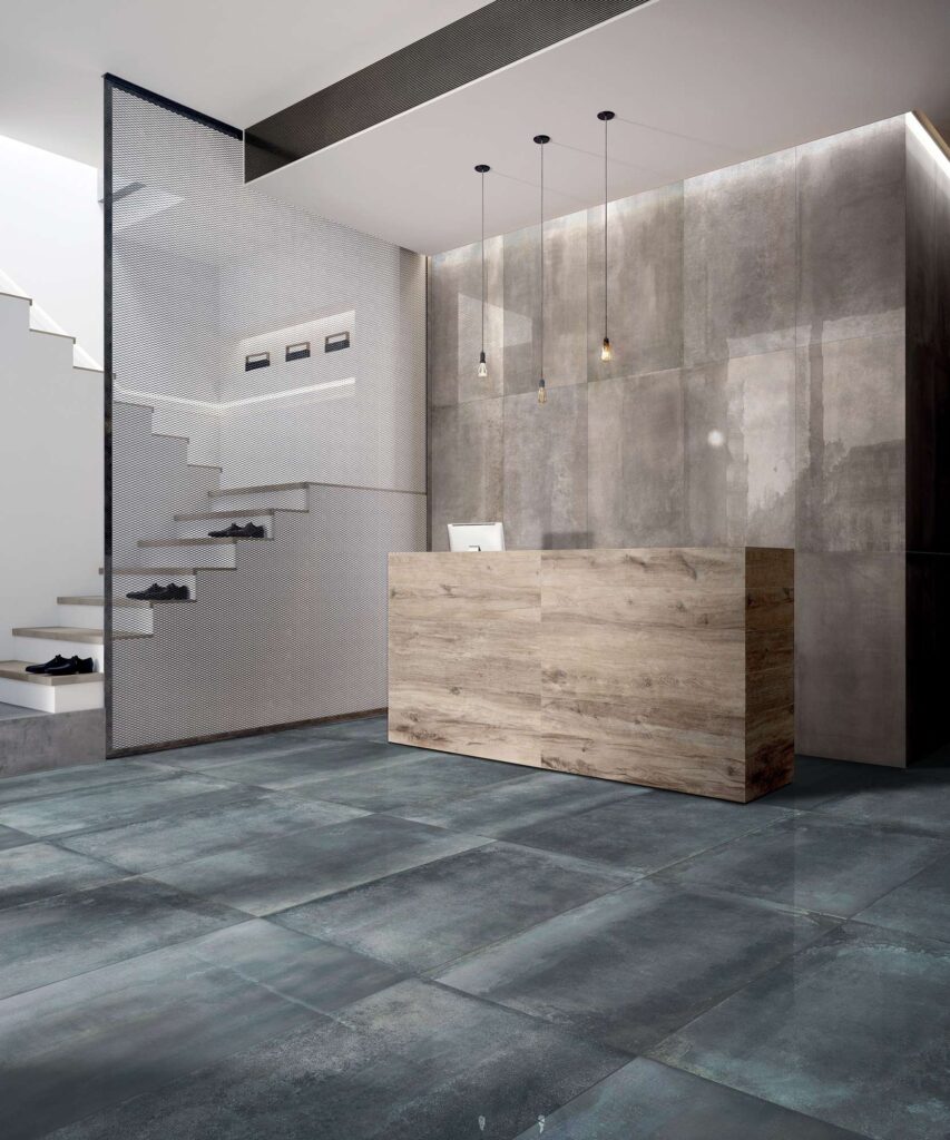 La collezione HEMISPHERE di gres porcellanato, proposta nei colori Lagune e Steel nel formato 60x120 cm con finitura lappata, definisce lo stile di questa elegante e moderna reception. Il pavimento è caratterizzato dalle piastrelle effetto metallo cangiante nella sfumatura verde-azzurra Lagune, mentre la parete di fondo è rivestita con la variante grigia Steel. L'ambiente, dall'atmosfera contemporanea e professionale, presenta un bancone in legno naturale, minimali lampade a sospensione e una scala di design separata da un divisore in rete metallica.