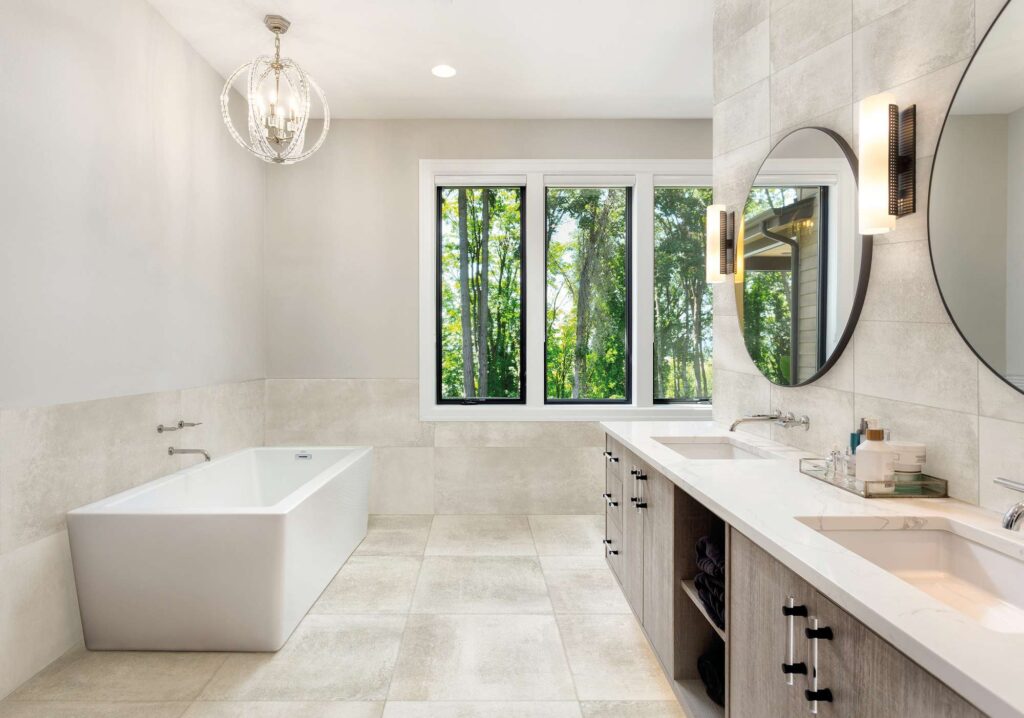 La collezione di piastrelle Hemisphere colore Platinum arreda un bagno moderno e luminoso, mostrando la versatilità dei formati 60x60 e 60x120. A pavimento, le piastrelle 60x60 in gres porcellanato grigio chiaro creano una superficie elegante e materica, mentre il formato 60x120 riveste parzialmente le pareti e la zona retrostante la vasca da bagno freestanding bianca. L'ambiente, caratterizzato da un mobile lavabo sospeso in legno con top chiaro, specchi circolari e una grande finestra affacciata sulla natura, esprime un'atmosfera di raffinato benessere e design contemporaneo.