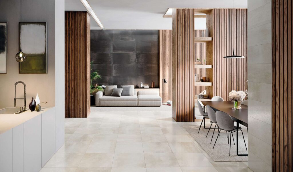 La collezione di pavimenti e rivestimenti Hemisphere arreda un elegante open space living, combinando armoniosamente le varianti Platinum e Iron. A pavimento, le piastrelle ceramiche chiare Hemisphere Platinum nel formato 60x60 cm donano luminosità e continuità visiva alla zona pranzo e all'area cucina. Sullo sfondo, la parete della zona giorno è valorizzata dal rivestimento scuro effetto metallo ossidato Hemisphere Iron nel formato 60x120 cm, posato in orizzontale. L'ambiente, dal design sofisticato e contemporaneo, è arricchito da quinte architettoniche con listelli verticali in legno, un moderno tavolo da pranzo con sedie grigie e un accogliente divano in tessuto chiaro, creando un'atmosfera calda e lussuosa.