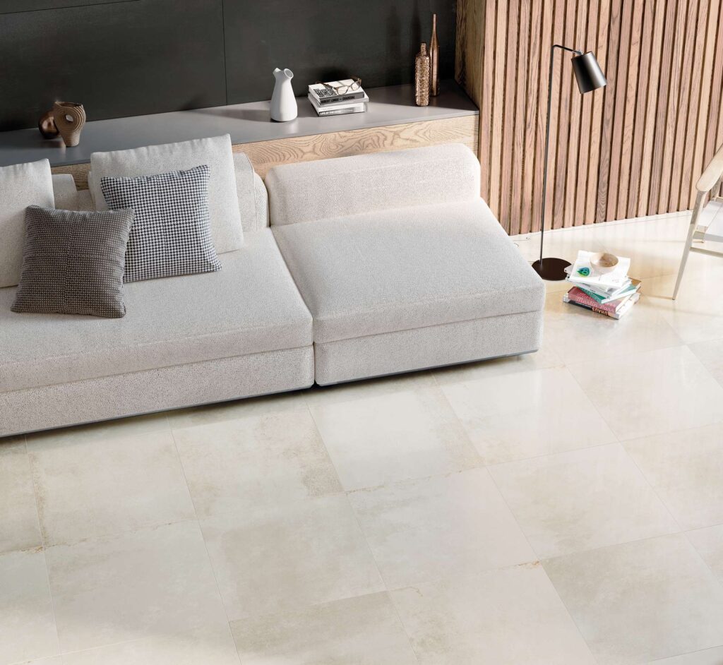 La collezione Hemisphere in gres porcellanato, proposta nelle varianti Platinum formato 60x60 cm e Iron formato 60x120 cm, veste con eleganza un moderno ambiente living di design. A pavimento, la posa regolare delle piastrelle chiare dal finish luminoso Platinum 60x60 esalta la luminosità della stanza, dialogando armoniosamente con un divano modulare color panna arricchito da cuscini decorativi. Sullo sfondo, la parete scura rivestita con le lastre Iron 60x120 e i dettagli in legno cannettato creano un contrasto materico sofisticato, offrendo un'atmosfera calda, lussuosa e accogliente.