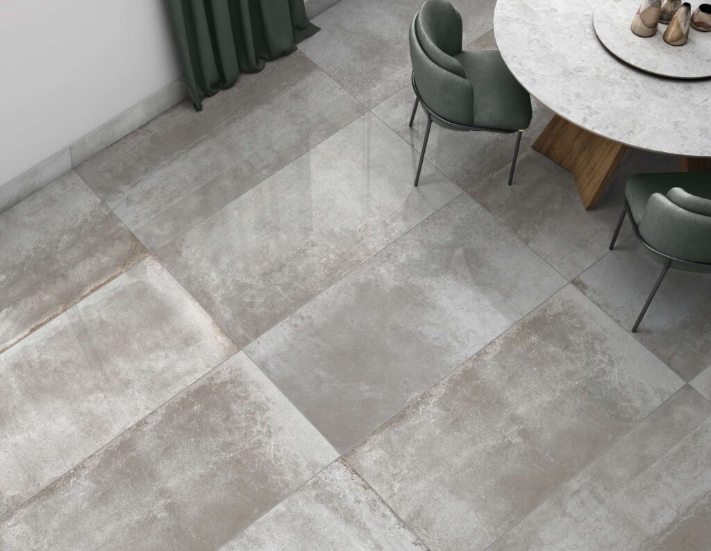 La collezione Hemisphere, nella variante colore Steel in formato 60x120 cm, riveste con eleganza il pavimento di una raffinata zona pranzo. Le grandi lastre in gres porcellanato effetto metallo grigio, caratterizzate da sfumature materiche e una superficie con delicati riflessi di luce, sono posate in modo regolare. L'ambiente, dall'atmosfera sofisticata e contemporanea, è completato da un tavolo da pranzo rotondo con piano in marmo chiaro e gambe in legno, accostato a poltroncine di design in velluto verde salvia e a una tenda coordinata.