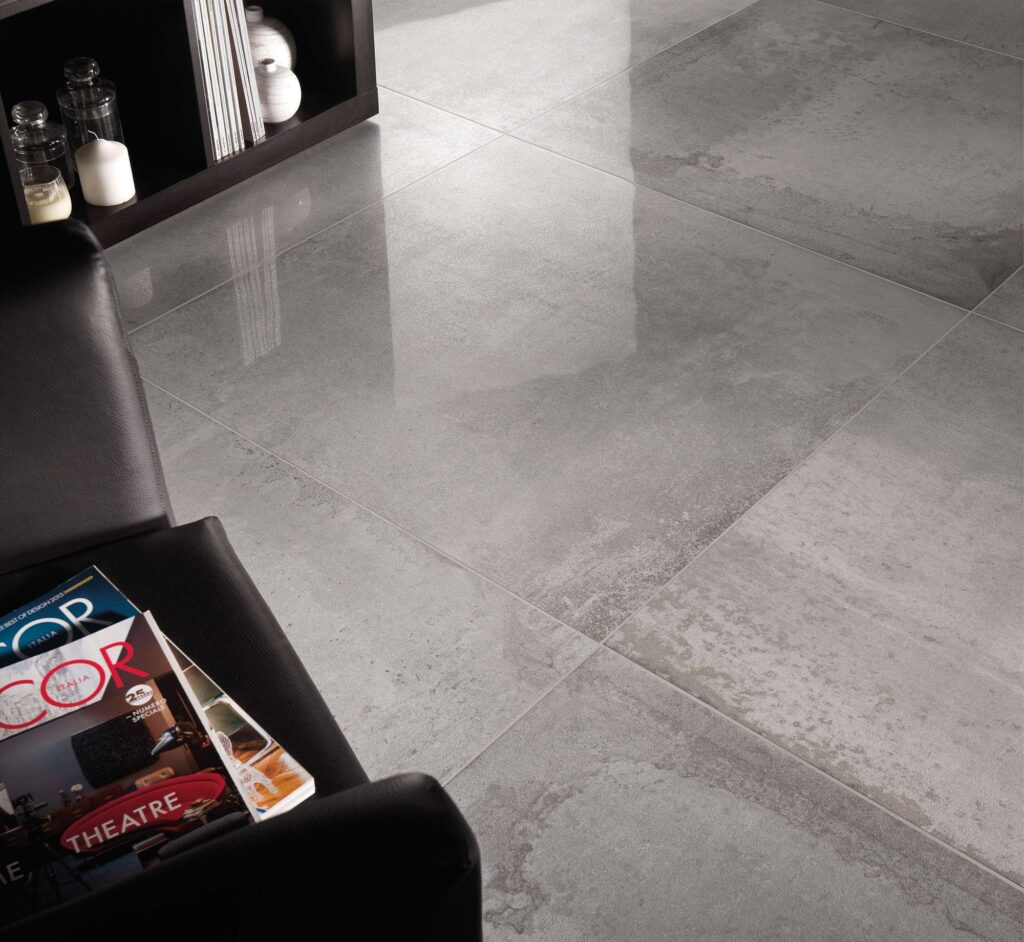 La collezione di piastrelle in gres porcellanato Hemisphere colore Steel nel formato 80x80 cm con finitura lappata è qui presentata in un'elegante posa a pavimento per interni. L'inquadratura ravvicinata valorizza la superficie grigia effetto metallo e la sua texture semilucida che riflette delicatamente la luce. L'ambiente, dallo stile moderno e sofisticato, è arricchito da una poltrona in pelle nera con riviste in primo piano e da un mobile di design scuro con oggetti decorativi.